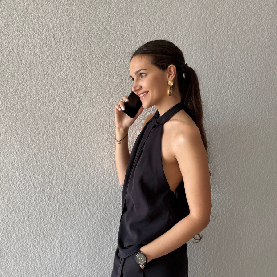 Mujer joven hablando por teléfono, vestida de negro, sonriente, de perfil, junto a una pared clara